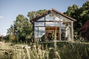 Een modern glazen huis met een schuin dak, omringd door hoog gras en bomen.