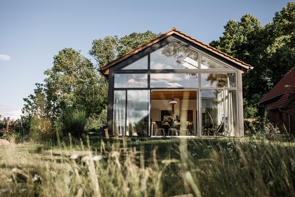 Una casa moderna de vidrio con techo inclinado, rodeada de pastos altos y árboles.