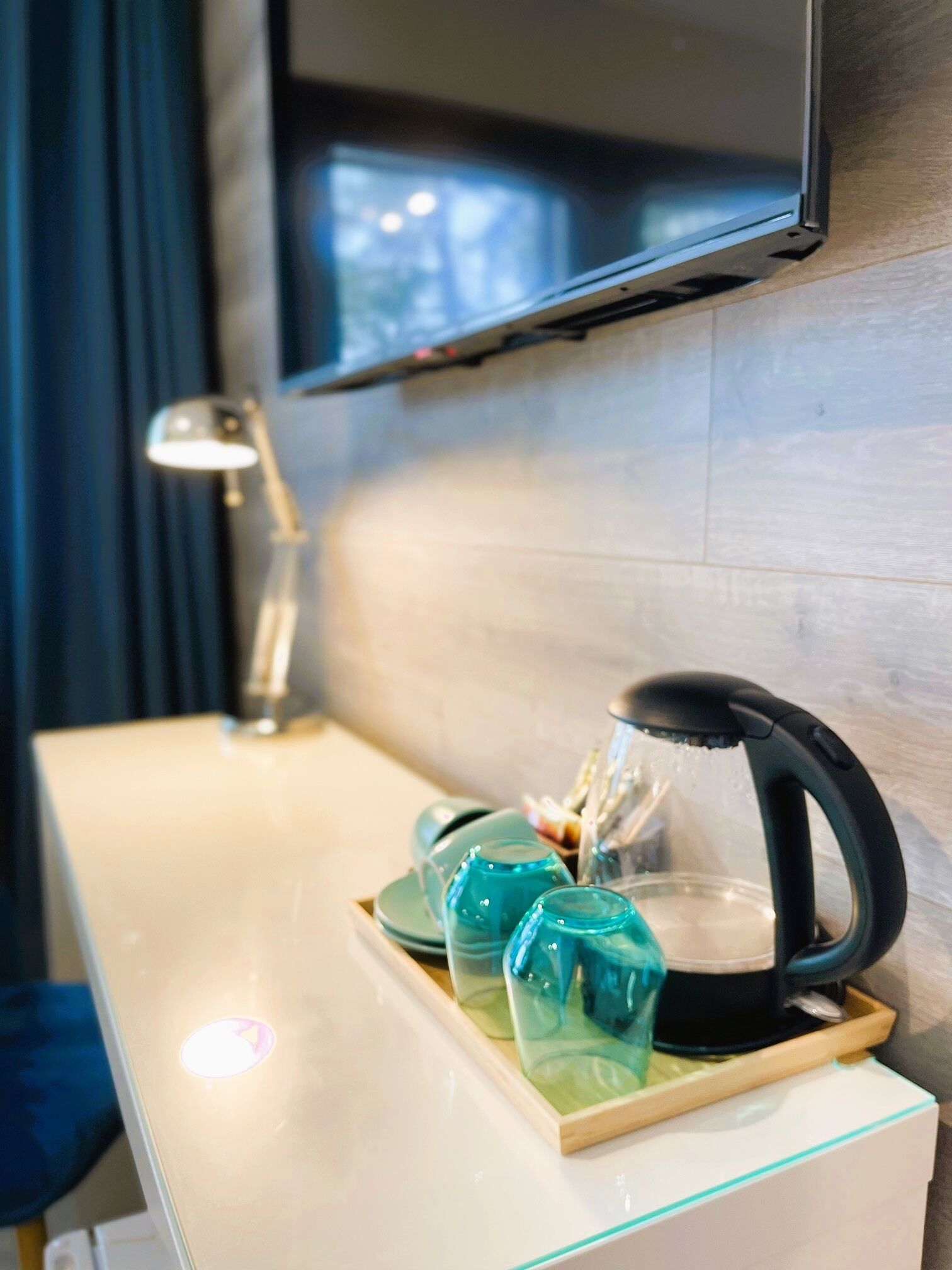 A hotel room with a wooden desk, a black kettle, and teal glass tumblers on a tray.