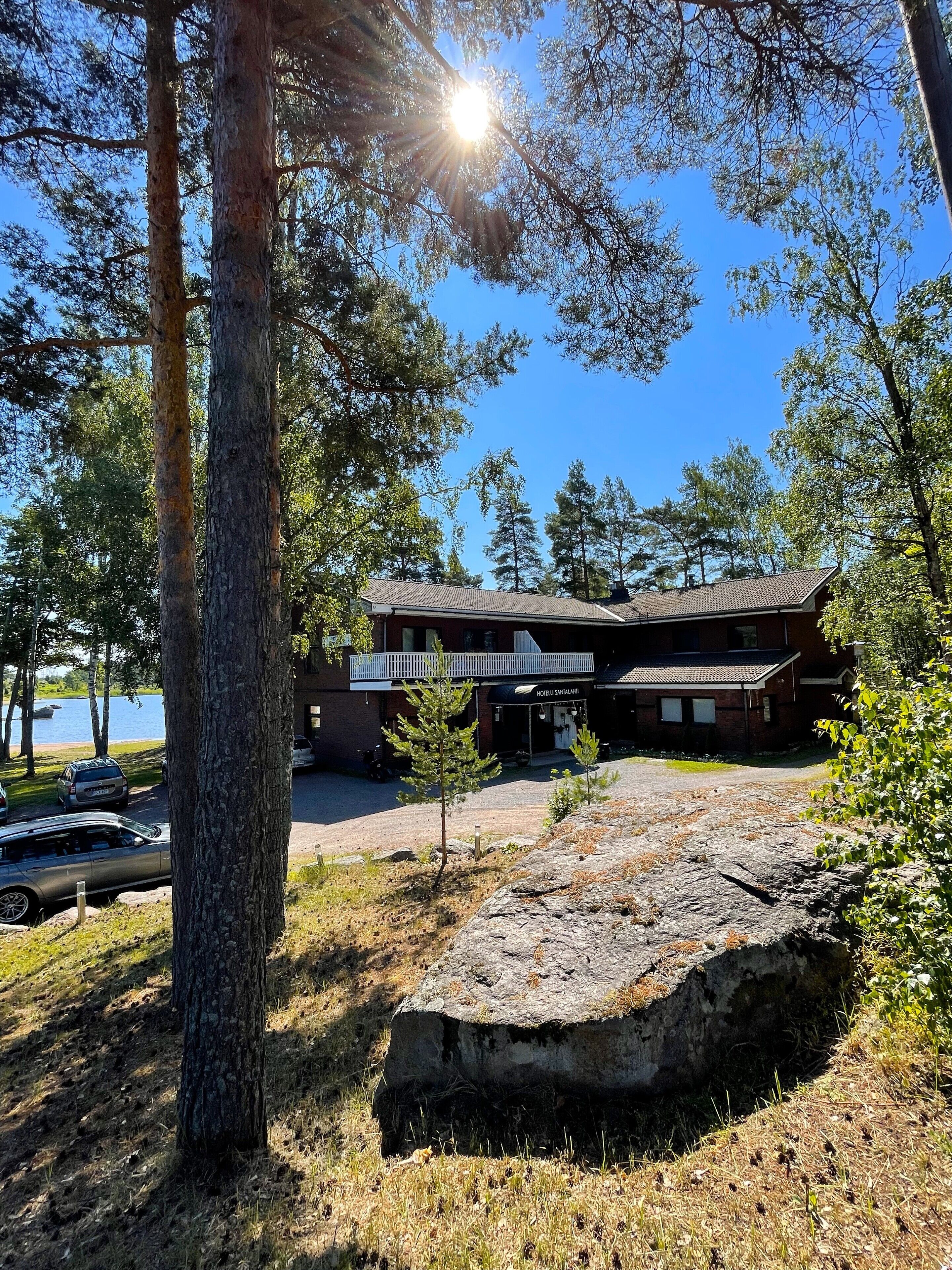 A lodge with a large rock in the foreground, a parking area with cars, and a view of the lake.