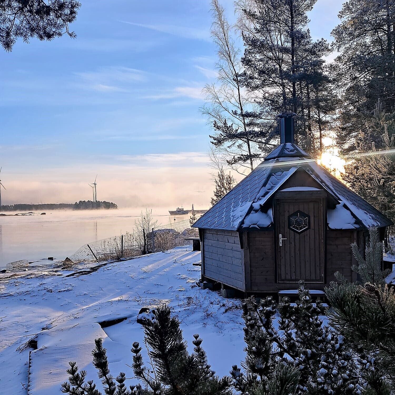 A wooden cabin with a snow-covered roof, situated by a frozen lake with a view of a distant cityscape and a ship in the background.