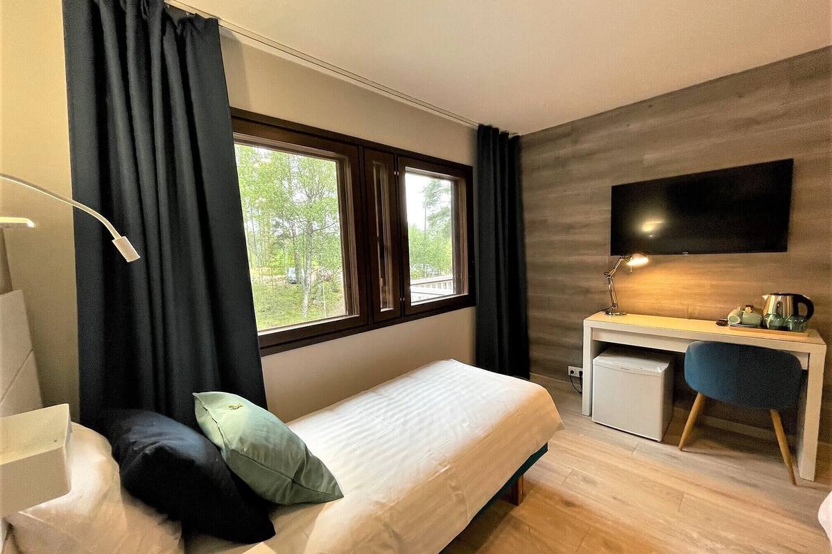 A bedroom with a bed, a desk, a chair, a TV, and a window with curtains.