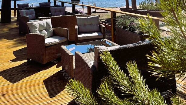 A wooden deck with wicker furniture and a glass table, overlooking a body of water with a distant wind turbine.