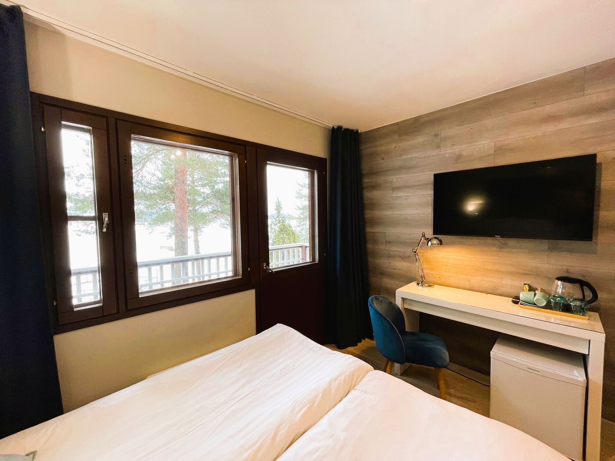 A hotel room with a bed, a desk, a chair, a TV, and a window with a view of trees and a balcony.