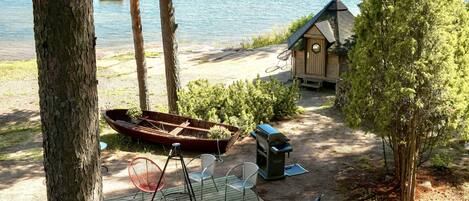 A lakeside cabin with a boat, outdoor seating, and a small shed.