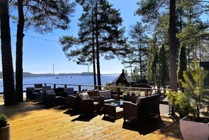 A deck with outdoor furniture overlooking a body of water, with trees and a small building in the background. - Beach Hotel Santalahti (Kotka)