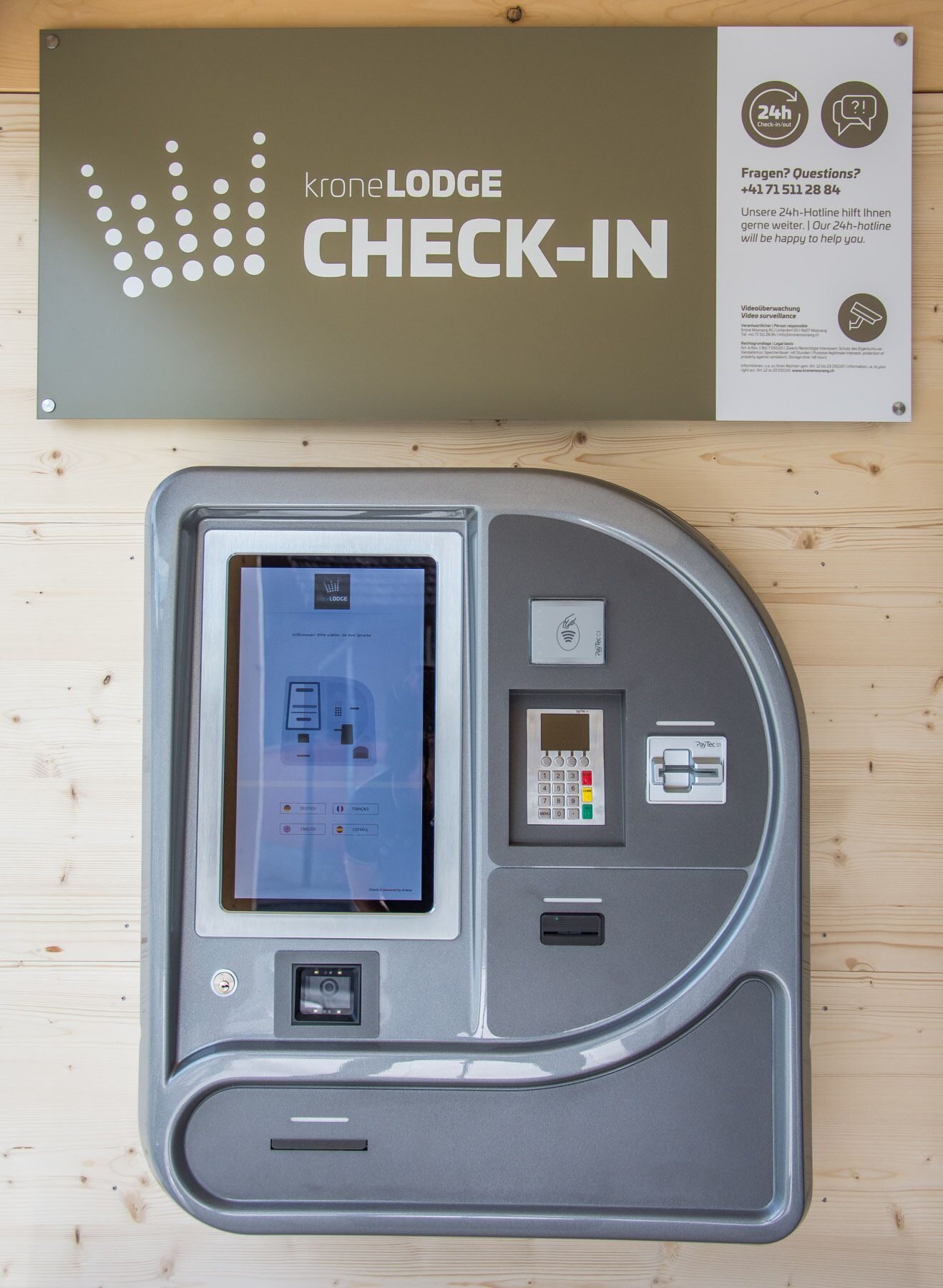 A check-in kiosk with a screen, keypad, and card reader, mounted on a wooden wall.