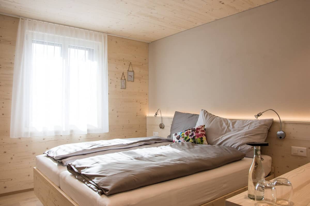 Un dormitorio con techo de madera, una cama con dos almohadas, una mesita de noche con una botella y vasos, y una ventana con cortinas de encaje.