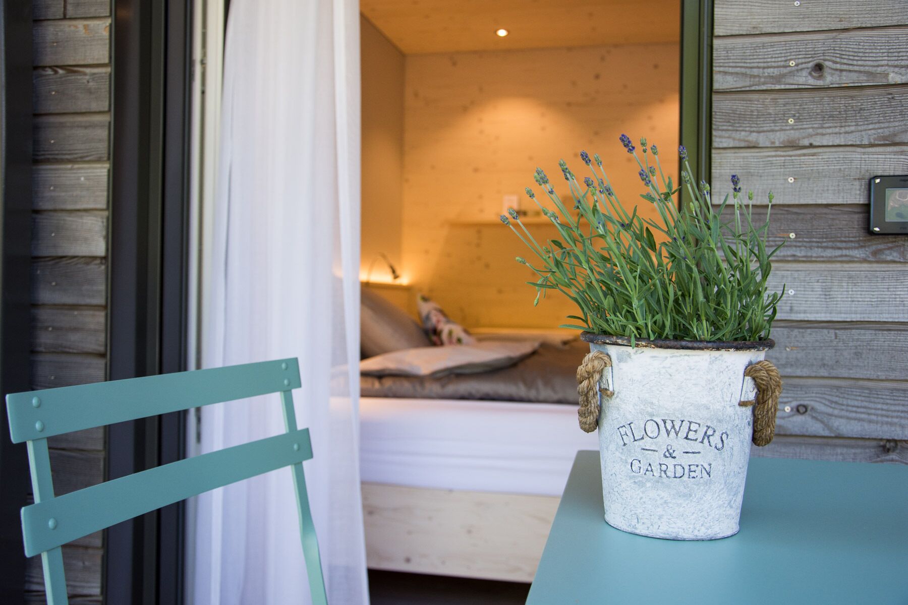Una habitación rústica con una cama, una mesita con una maceta de lavanda y una silla verde.