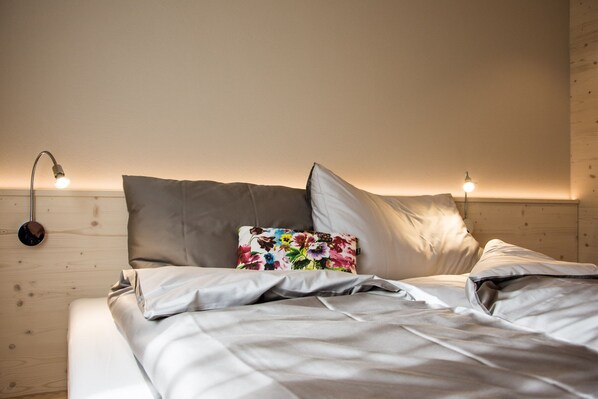 A neatly made bed with white linens, a colorful floral pillow, and two bedside lamps on a wooden wall. - kroneLODGE (Mosnang)