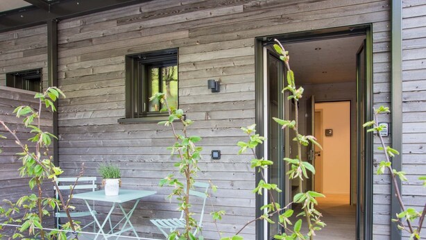 Un edificio de madera con puerta de vidrio, una pequeña mesa con sillas y una planta en maceta.