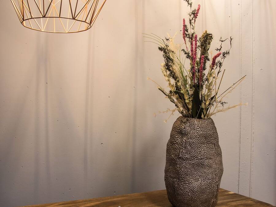 A wooden table with a textured vase containing dried flowers, two geometric pendant lights, and a stack of magazines.