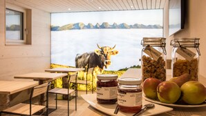 Un comedor con mesas y sillas de madera, un ventanal con vistas a una vaca y montañas, y un recipiente con mermelada y manzanas.