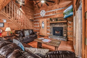 L’intérieur d’une cabane en rondins, avec une cheminée, des canapés en cuir, un escalier en bois et une horloge au mur.