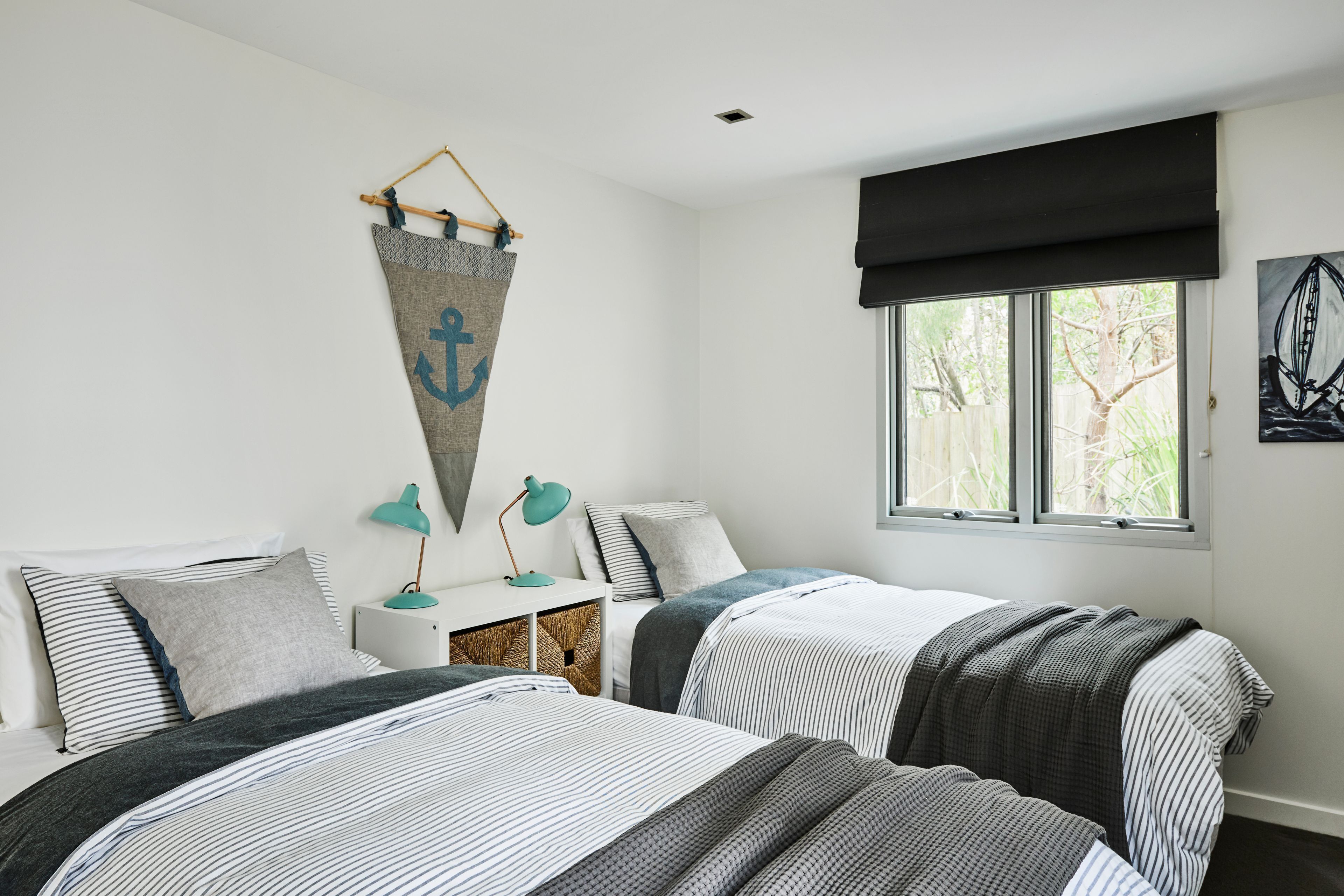 A bedroom with two beds, a window with a view of trees, a wall-mounted anchor decoration, and a desk with a lamp.