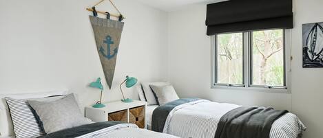 A bedroom with two beds, a window with a view of trees, a wall-mounted anchor decoration, and a desk with a lamp.