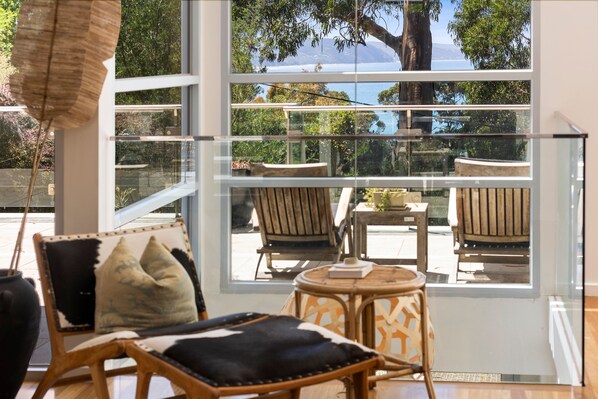 A room with a large window offering a view of the sea and trees. There are two wooden chairs and a small round table with a book on it.