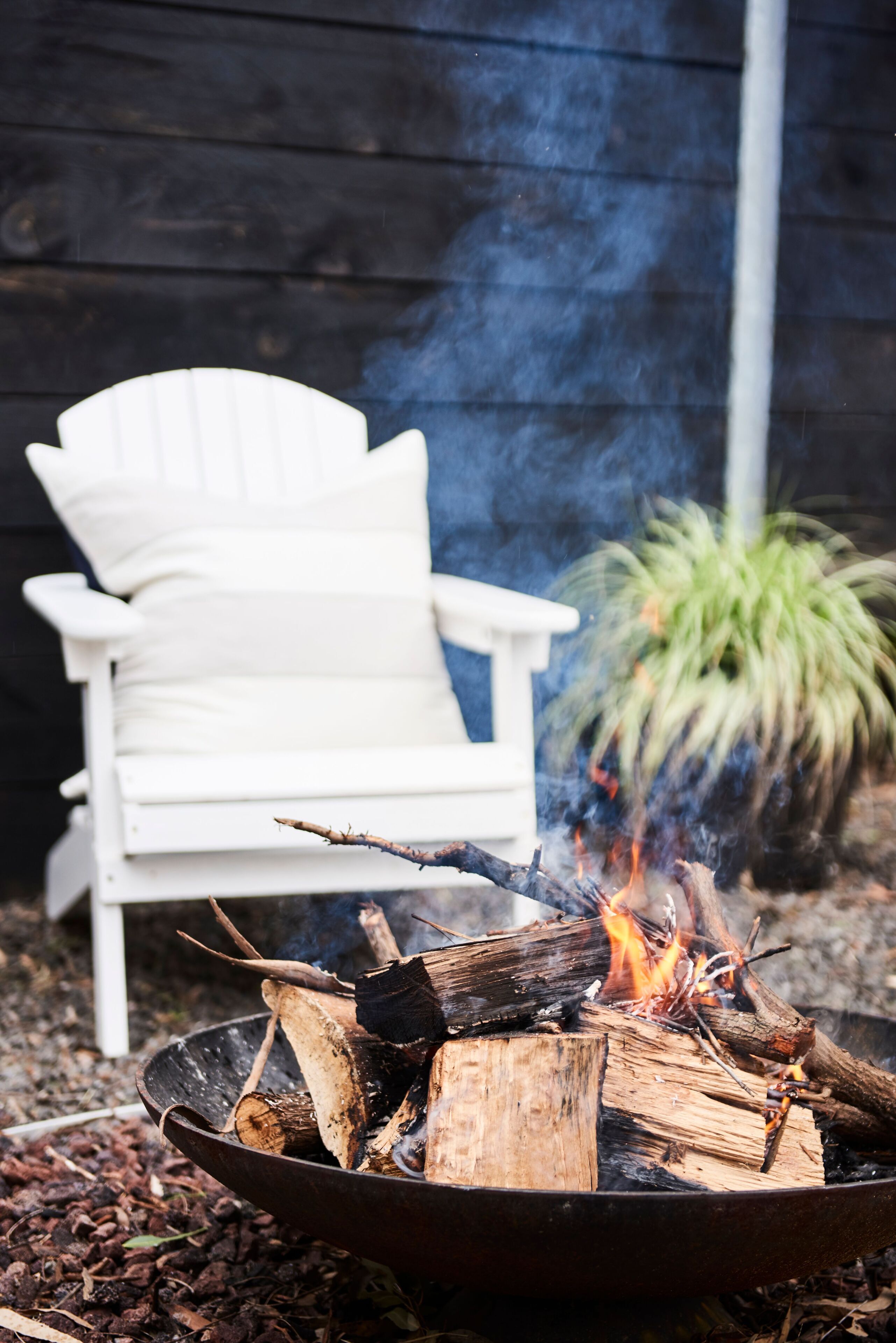 A campfire with logs and embers, a white chair, and a potted plant in the background.