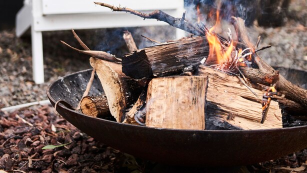 A campfire with logs and embers, a white chair, and a potted plant in the background. - Seaview House Luxury Coastal Escape Lorne (Lorne)