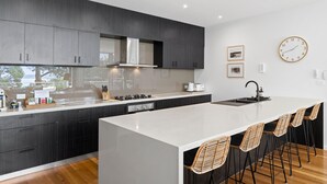 A modern kitchen with dark cabinetry, a white island, and a bar area with wicker chairs. - Seaview House Luxury Coastal Escape Lorne (Lorne)