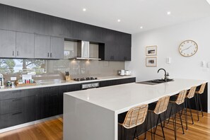 A modern kitchen with dark cabinetry, a white island, and a bar area with wicker chairs. - Seaview House Luxury Coastal Escape Lorne (Lorne)