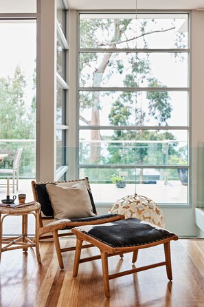 Ein Zimmer mit großem Fenster, einem hölzernen Stuhl, einem kleinen Tisch und einem gepolsterten Hocker.