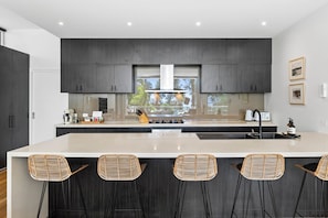 A modern kitchen with dark cabinetry, a central island with bar stools, and a view of the outdoors through large windows.