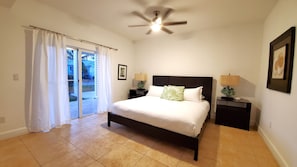 Une chambre à coucher avec un lit, deux tables de chevet, un ventilateur de plafond et une vue sur l’extérieur, accessible par une porte-fenêtre coulissante.