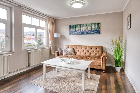 A living room with a beige sofa, a white coffee table, and a painting of a forest path on the wall.