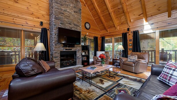 L’intérieur d’un chalet confortable, avec des murs en rondins, une cheminée en pierre, des canapés en cuir et une grande fenêtre équipée de stores.