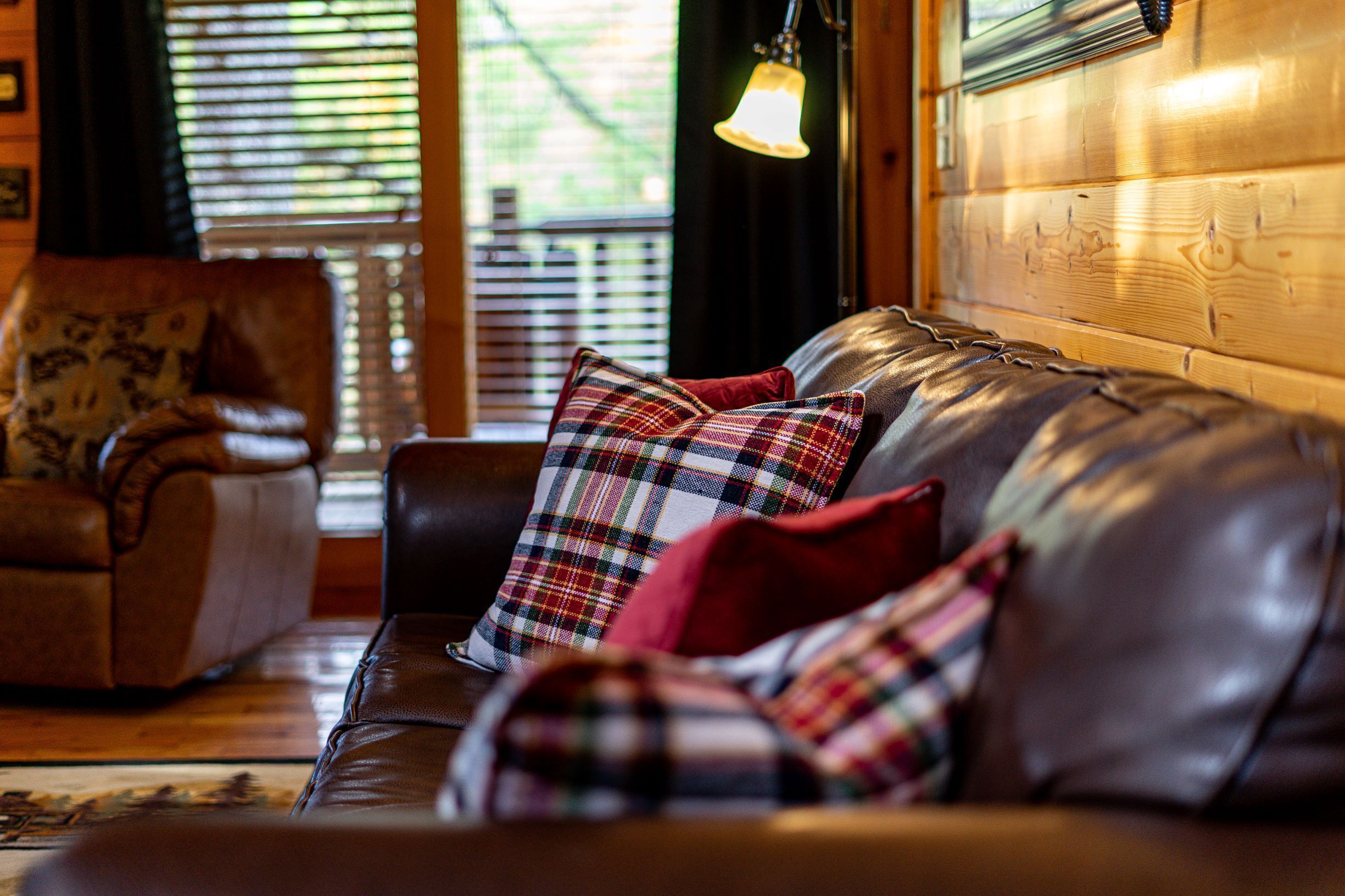 A leather sofa with plaid cushions in a room with wooden paneling and a window with blinds.