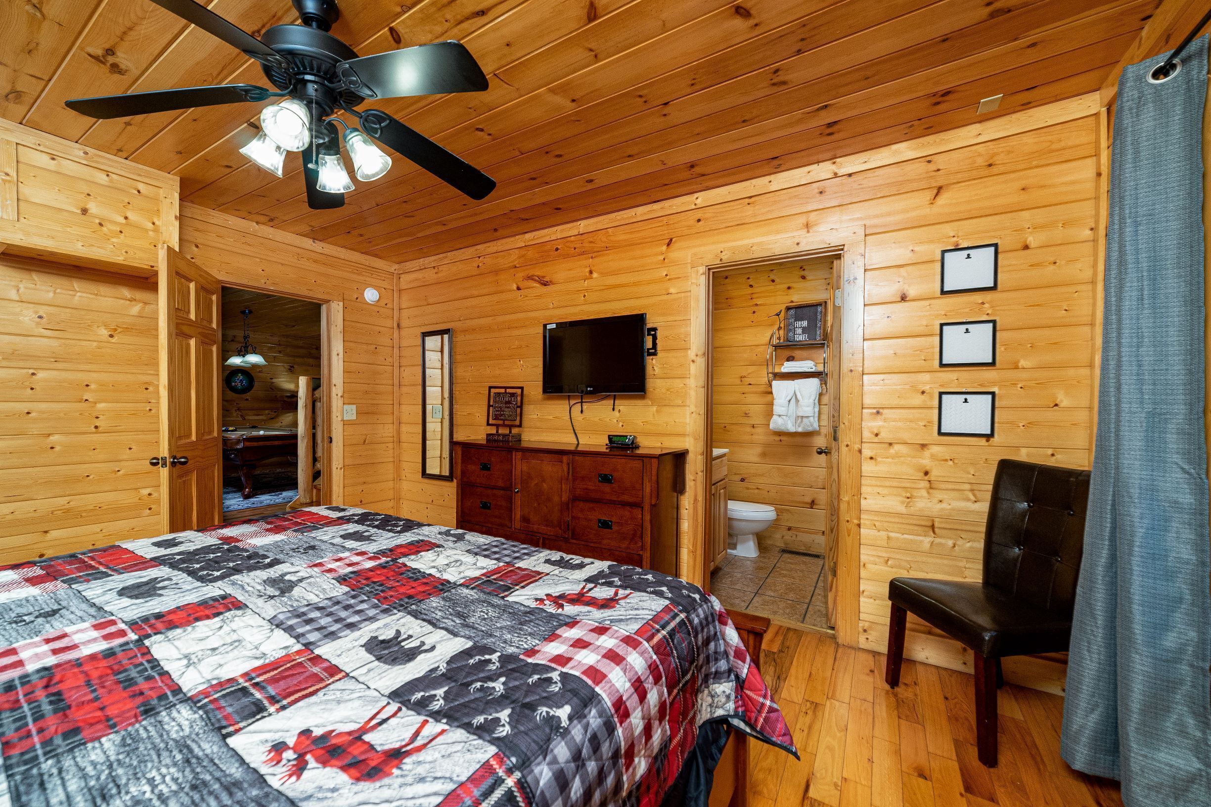 A wooden cabin bedroom with a bed, a ceiling fan, a television, and a wooden dresser.