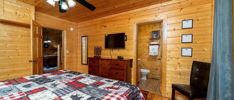 A wooden cabin bedroom with a bed, a ceiling fan, a television, and a wooden dresser.