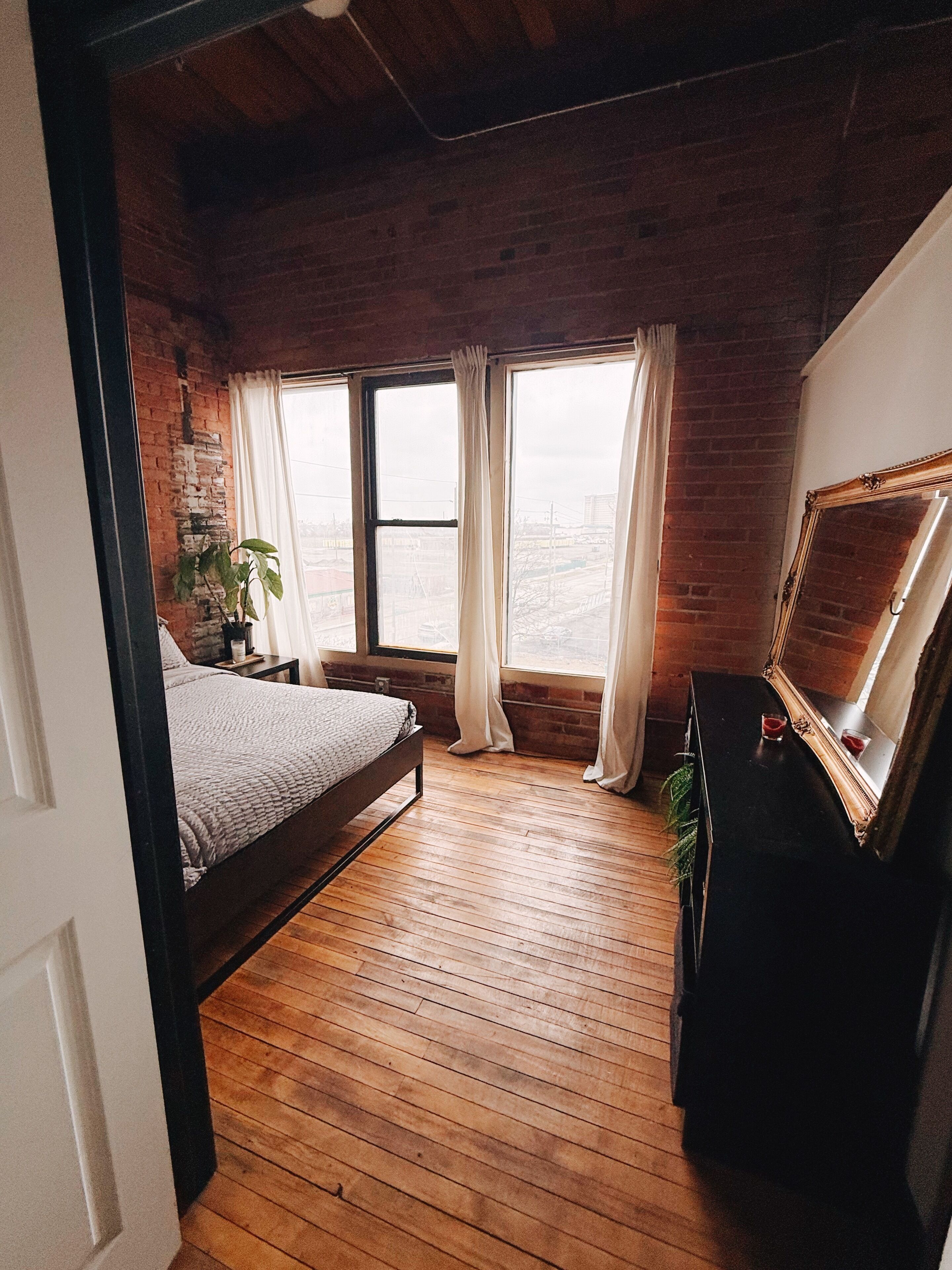 Una camera da letto con pavimento in legno, un letto, una cassettiera, uno specchio e una finestra con tende.