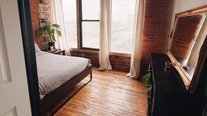 A bedroom with a wooden floor, a bed, a dresser, a mirror, and a window with curtains. - 420 Friendly Loft • Downtown Detroit Views, Walk to Riverfront, Free Parking (Detroit)