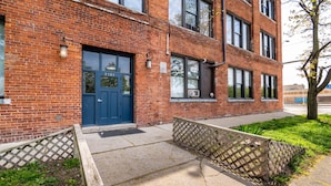 A brick building with a blue door, a small garden area with a wooden fence, and a tree with spring blossoms. - 420 Friendly Loft • Downtown Detroit Views, Walk to Riverfront, Free Parking (Detroit)