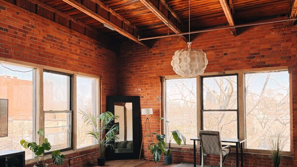 A spacious living room with a brick wall, large windows, a green sofa, a coffee table, and potted plants. - 420 Friendly Loft • Downtown Detroit Views, Walk to Riverfront, Free Parking (Detroit)