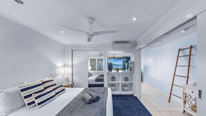 A modern bedroom with a bed, pillows, a bedside table, a nightstand, a ceiling fan, an air conditioning unit, a bookshelf, a ladder, and a view of the outside. - By The Sea Shore Stunning Unit Pool Spa Views (Cannonvale)