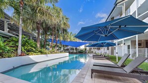 A poolside area with lounge chairs and umbrellas, surrounded by palm trees and a building with balconies. - By The Sea Shore Stunning Unit Pool Spa Views (Cannonvale)