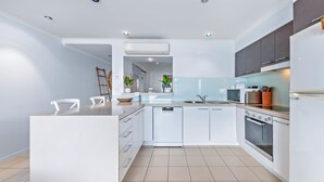 A modern kitchen with white cabinetry, a central island, and a built-in oven. - By The Sea Shore Stunning Unit Pool Spa Views (Cannonvale)