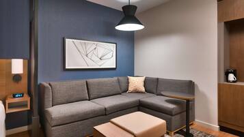 Una sala de estar moderna con un sofá seccional gris, una mesita redonda pequeña, una mesa de centro de madera, una obra de arte abstracto colgada en la pared y una lámpara colgante negra.