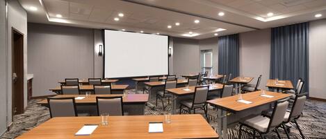 Une salle de conférence dotée de plusieurs tables et chaises, d’un grand écran de projection et d’un sol recouvert de moquette.