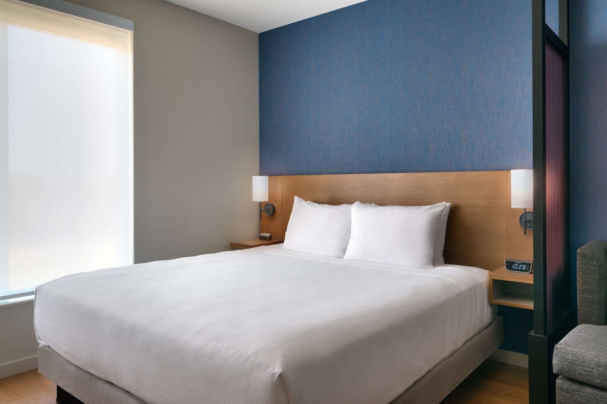 A hotel room with a large bed, two bedside tables, a nightstand, a clock, and a window with blinds.