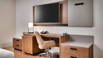 A modern hotel room with a wooden desk, a comfortable chair, a flat-screen TV, and a bedside table with drawers.