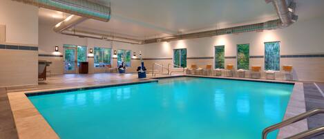 An indoor swimming pool with clear blue water, surrounded by lounge chairs and a poolside area with a view of trees.