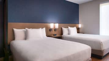 Habitación de hotel con dos camas, cada una con ropa de cama blanca y cabeceros. Hay mesitas de noche con lámparas y un reloj digital. El piso es de madera y una pared tiene detalles en azul.