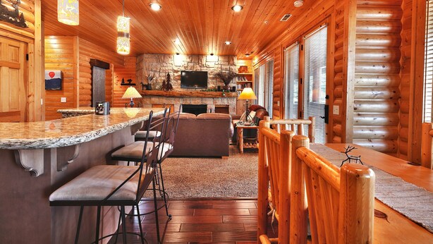 A log cabin interior with a stone fireplace, wooden bar, seating area, and a television mounted on the wall.