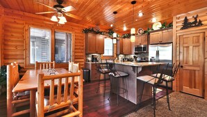 A wooden cabin interior with a dining table, chairs, a kitchen area, and a ceiling fan.