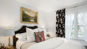 Cama bem arrumada com lençóis brancos e almofada estampada, mesa de cabeceira com abajur e um quadro de paisagem na parede.