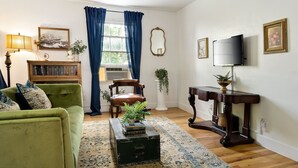 Uma sala de estar com um sofá verde, uma cadeira de madeira, uma mesinha com livros, uma cortina azul, um espelho e uma televisão na parede.